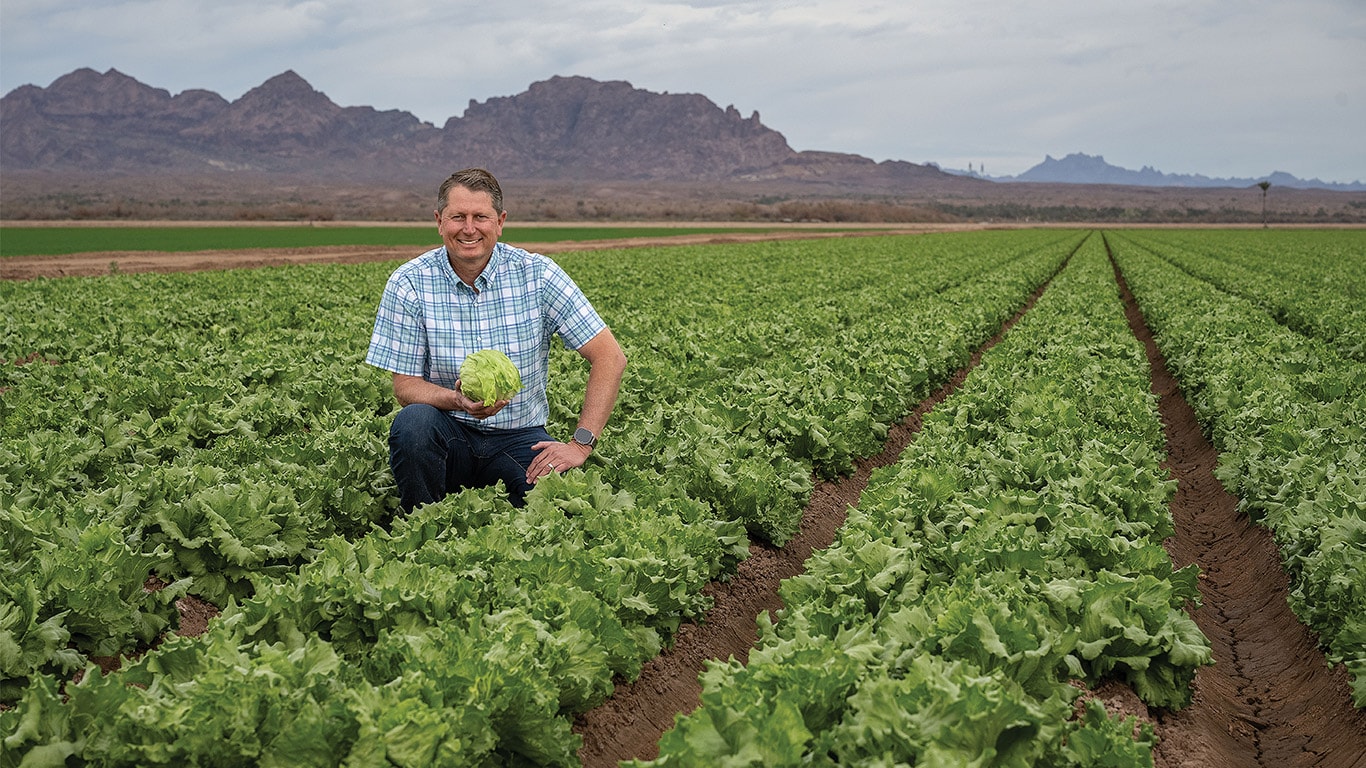 Individual kneeling down in a field of leafy greans while holding a head of lettuce.