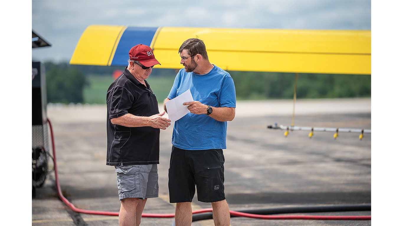 Two people review documents beside a crop-dusting aircraft on an airfield.