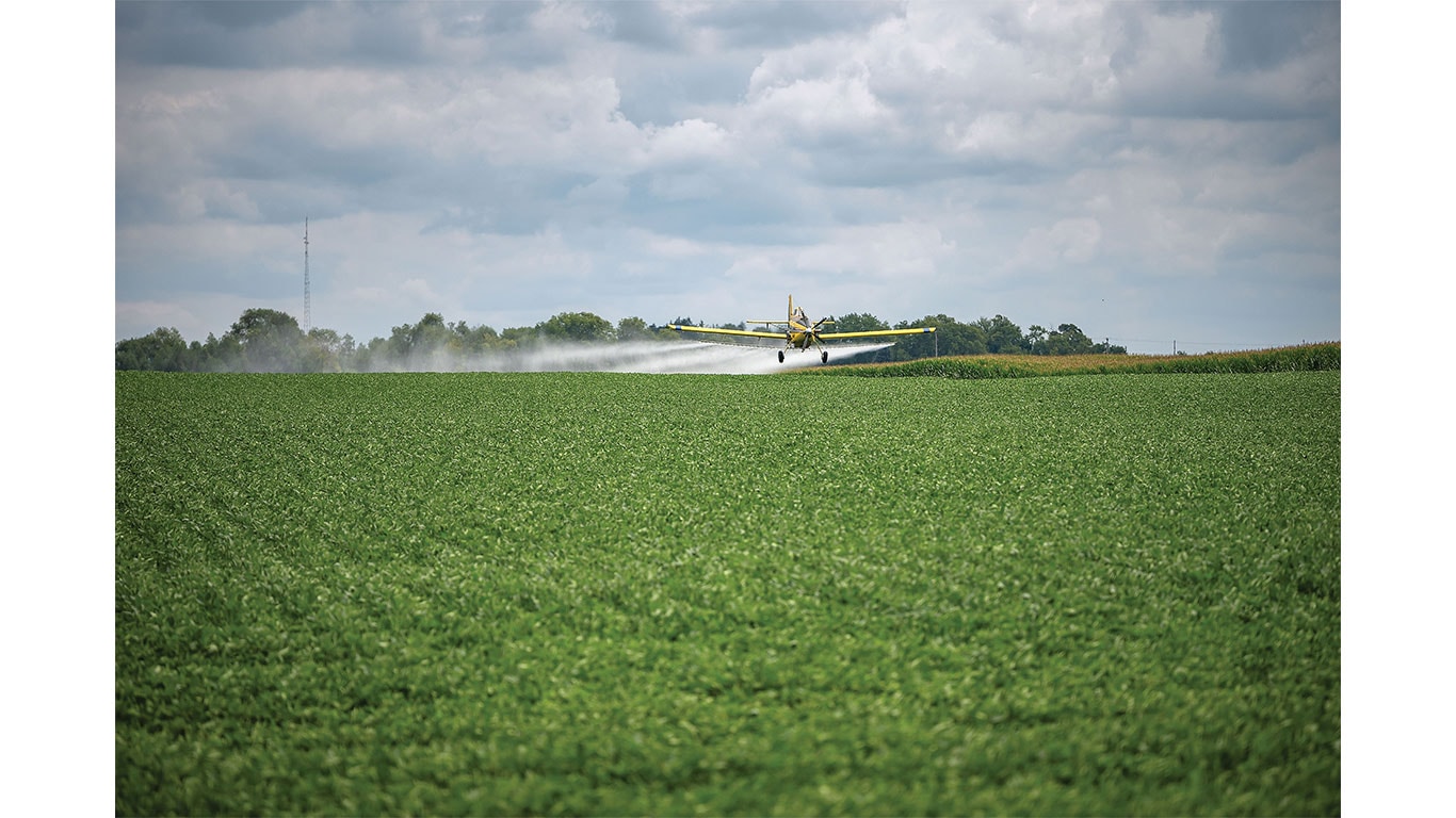 Crop-dusting aircraft sprays a green field beneath a cloudy sky.