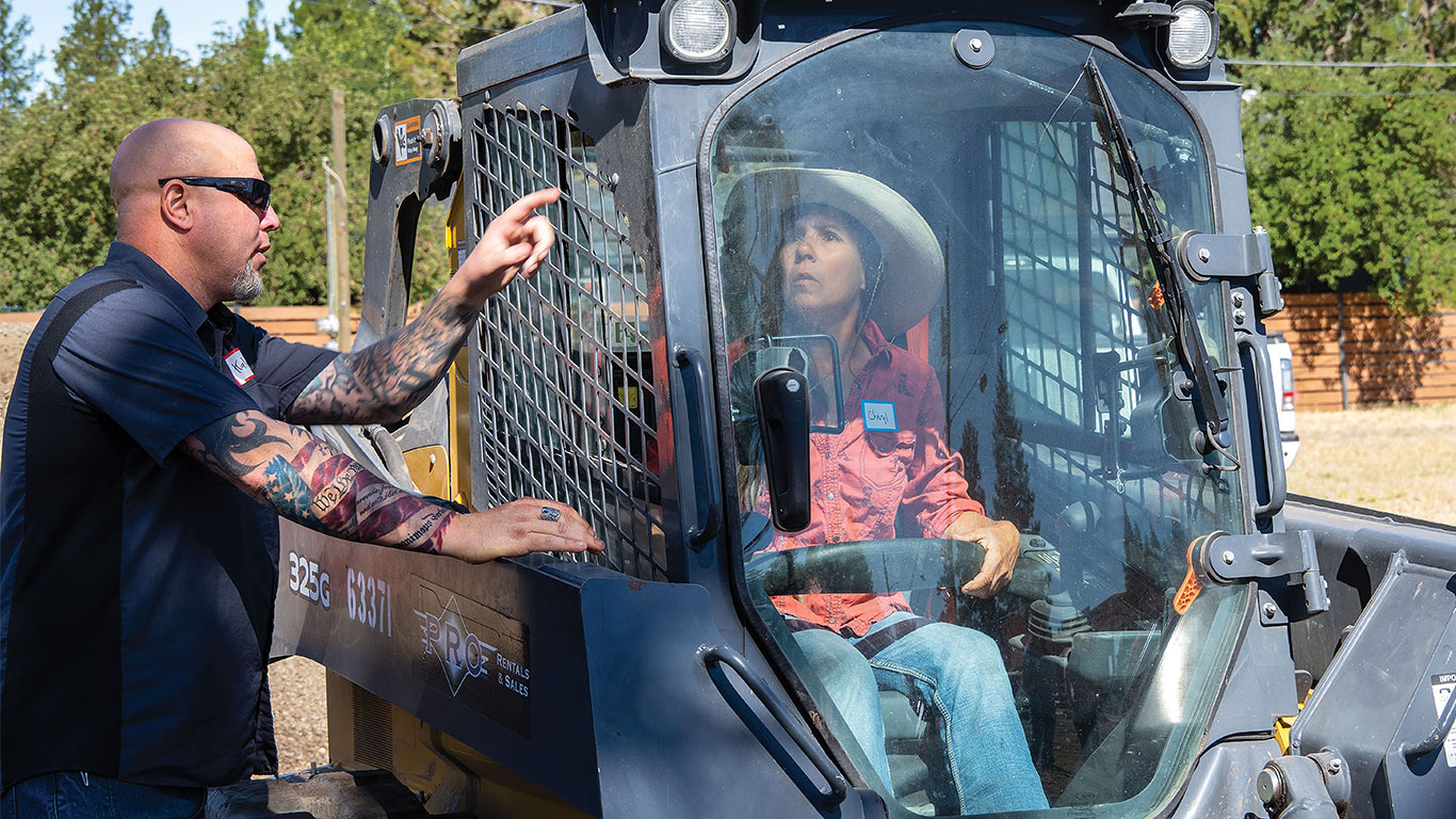 An instructor pointing to something in a skid steer with the rider looking at it too.