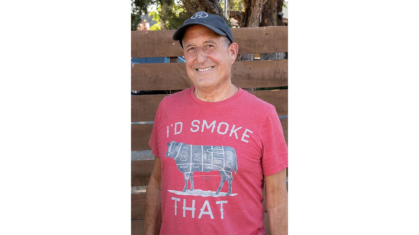 Person wearing a cap and graphic T-shirt standing in front of a wooden fence outdoors.