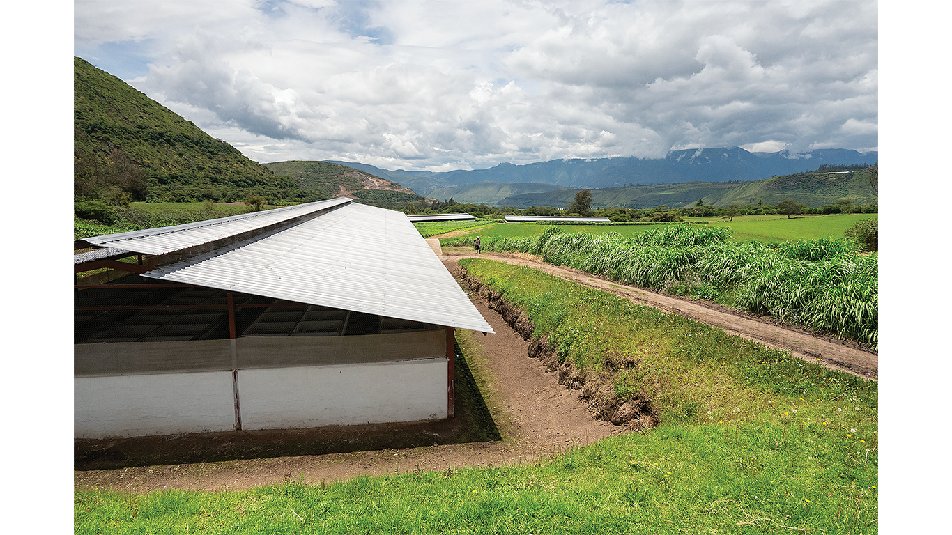 Farm building beside green fields with mountains and clouds in the background.
