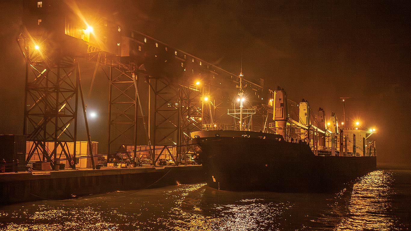 Cargo vessel moored at a lit industrial dock at night, with port cranes along the shoreline.