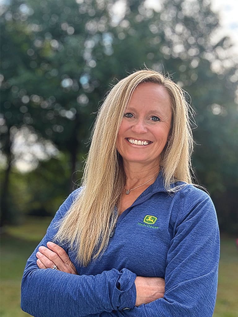 Person in a blue John Deere pullover standing outdoors with arms crossed.