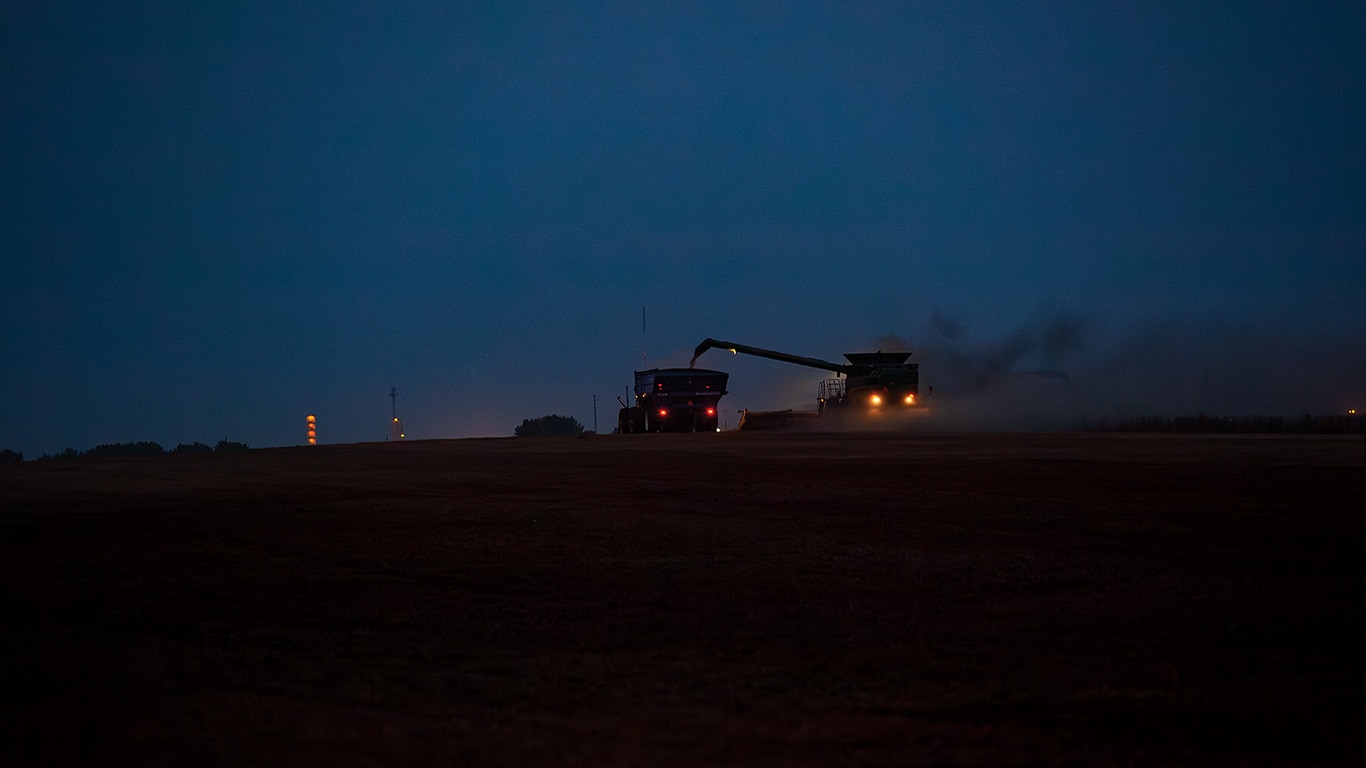 Combine and tractor harvesting in a dark field with headlights and dust in the air.
