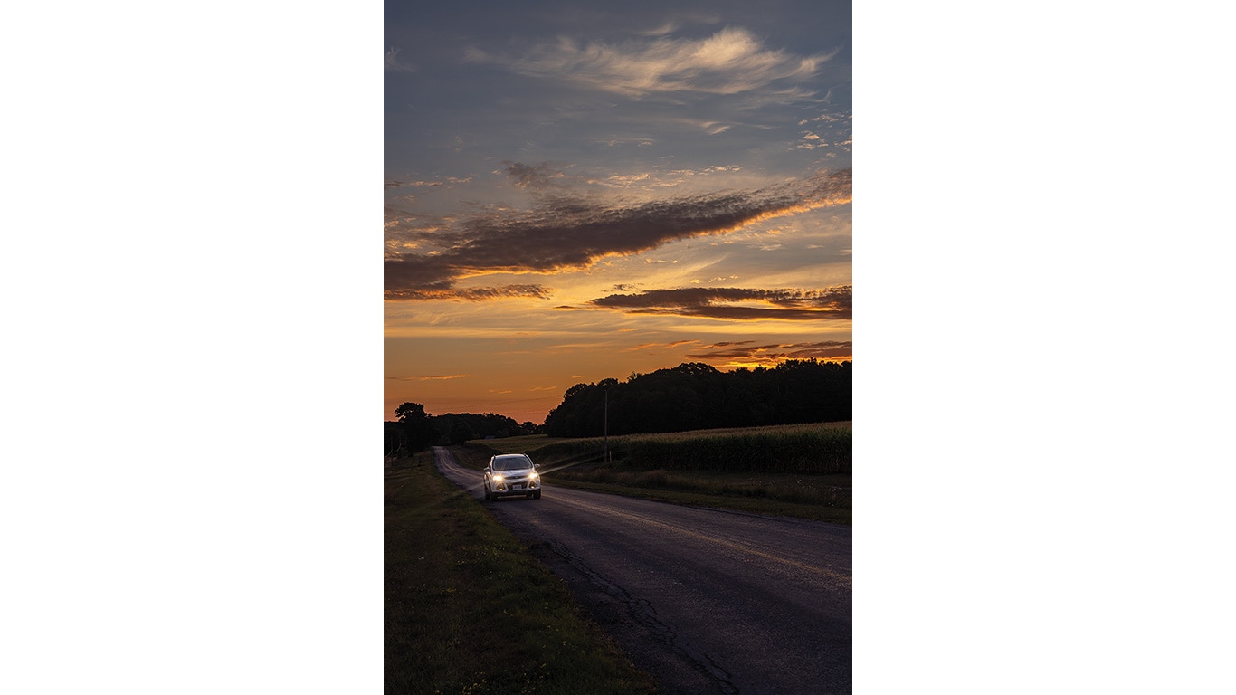 Car with headlights on driving down a rural road at sunset under a cloudy sky.