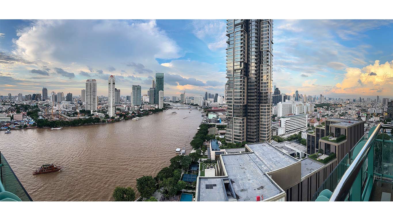 City skyline with tall buildings along a wide brown river under a partly cloudy sky.