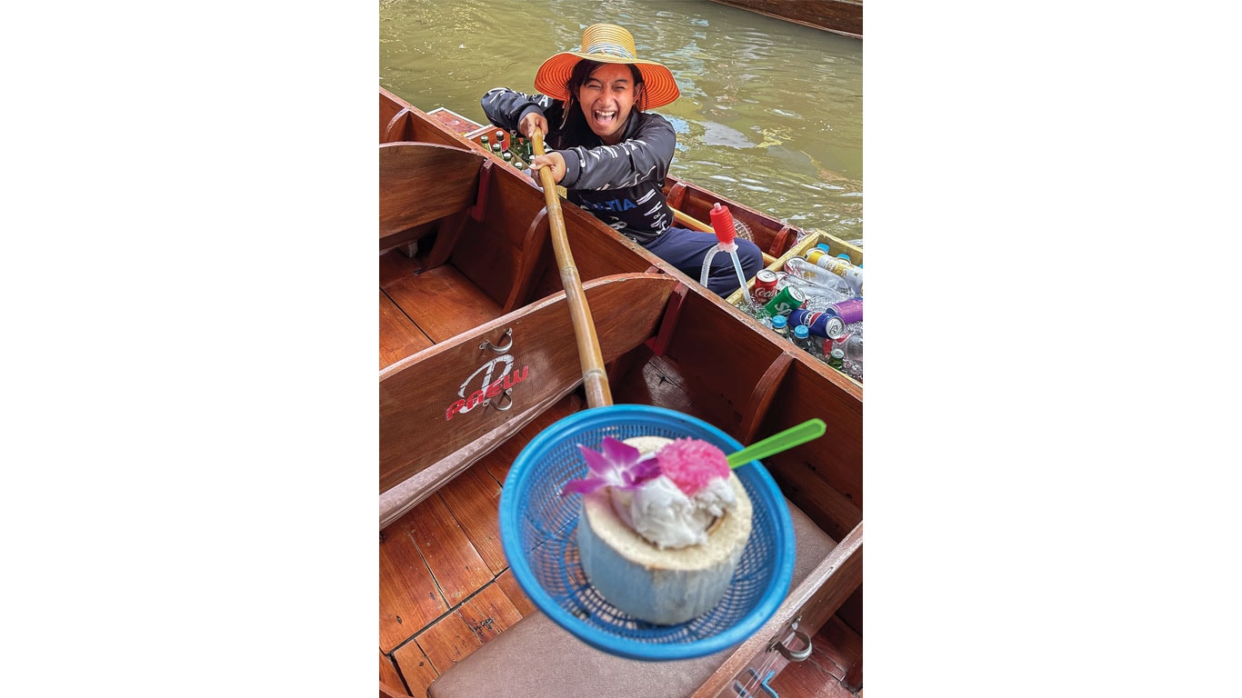 Coconut shell dessert with ice cream and orchid garnish held above wooden boats on a canal.