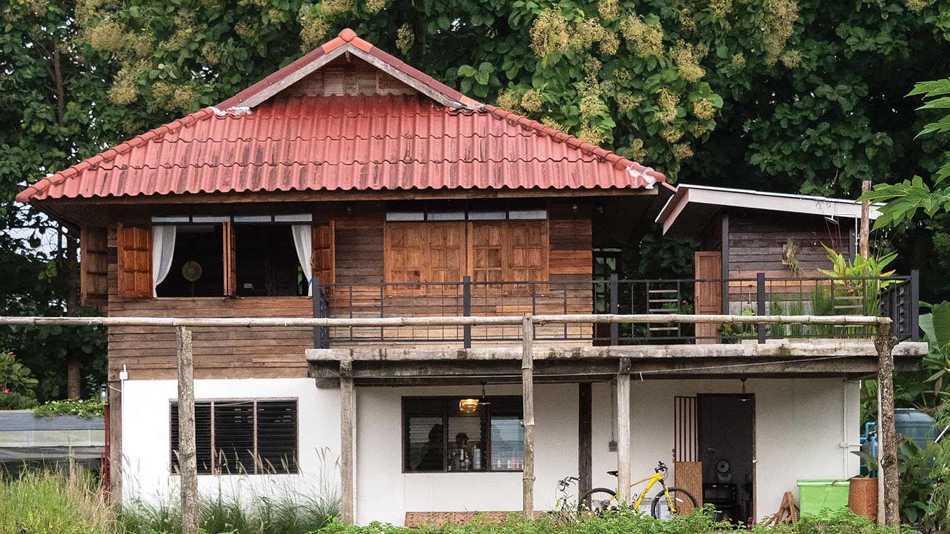 Two-story house with red tile roof, wooden upper walls, and white lower level surrounded by greenery.