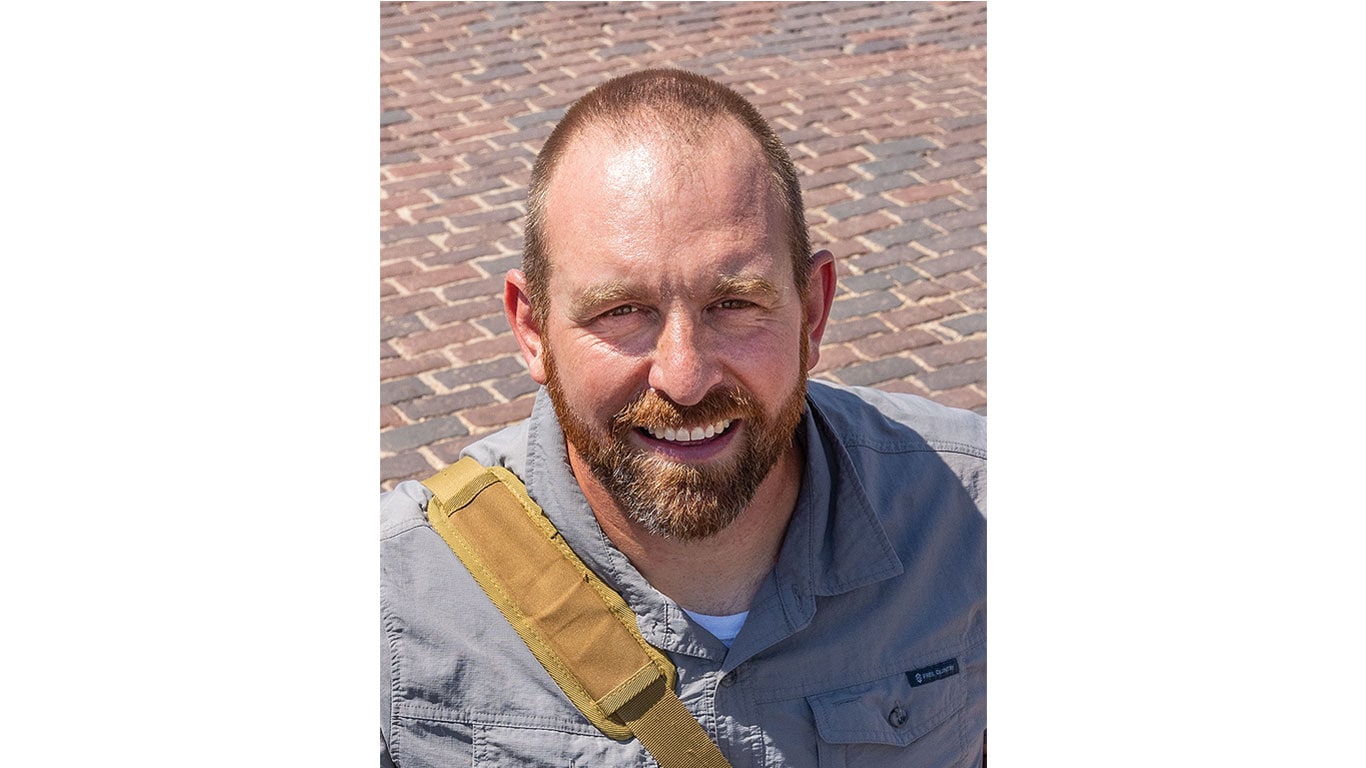 Person wearing a gray button-up shirt with a tan shoulder strap sitting on a cobblestone surface.