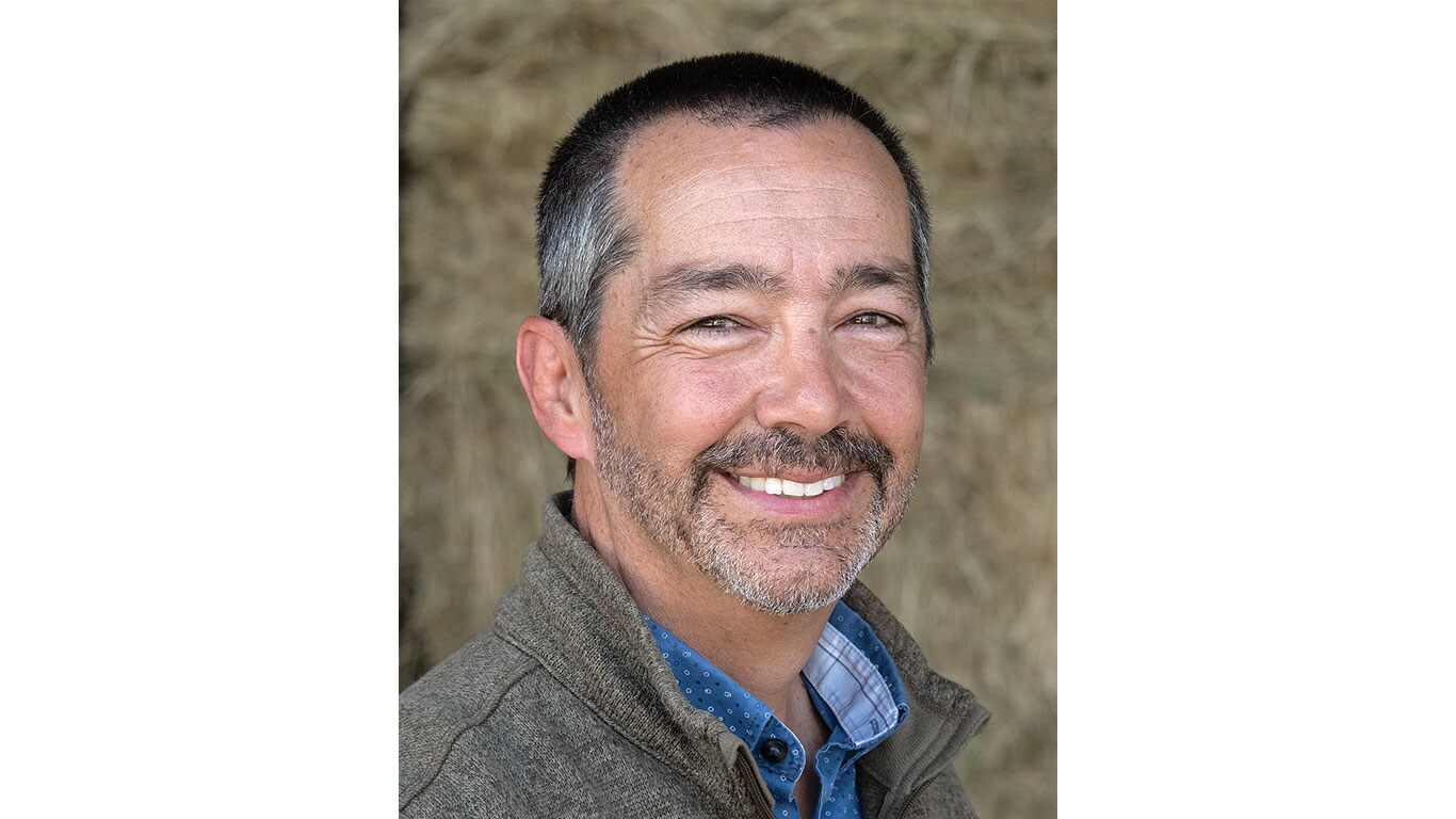Person wearing a gray zip-up jacket over a blue collared shirt standing in front of stacked hay.