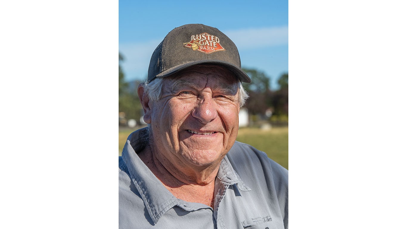 Person wearing a gray collared shirt and a dark cap with &ldquo;Rusted Gate Farm&rdquo; logo outdoors.