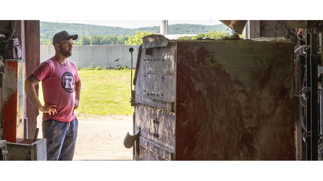 Person in a red shirt standing near a large metal feed mixer inside a barn with open doorway.