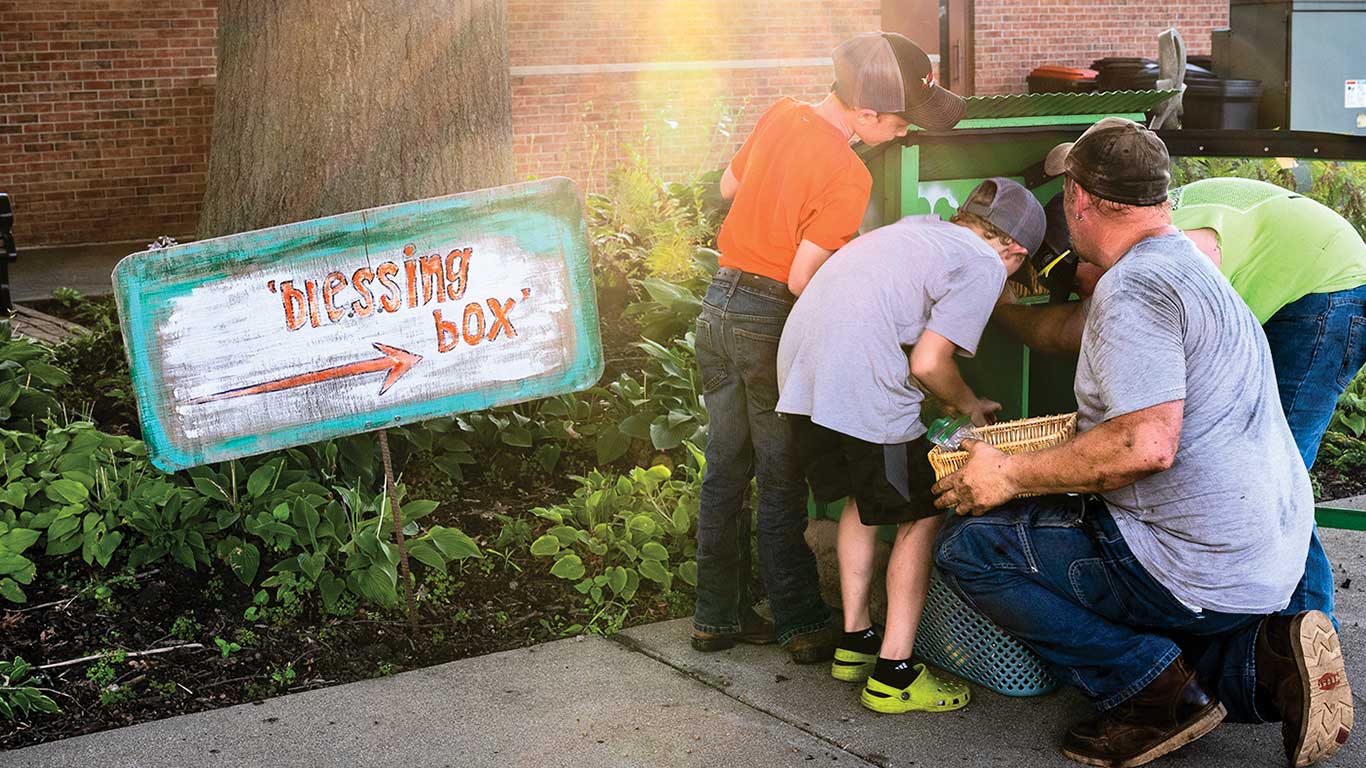 Adults and kids gather at a green outdoor box labeled 'Blessing Box' near a tree and plants.