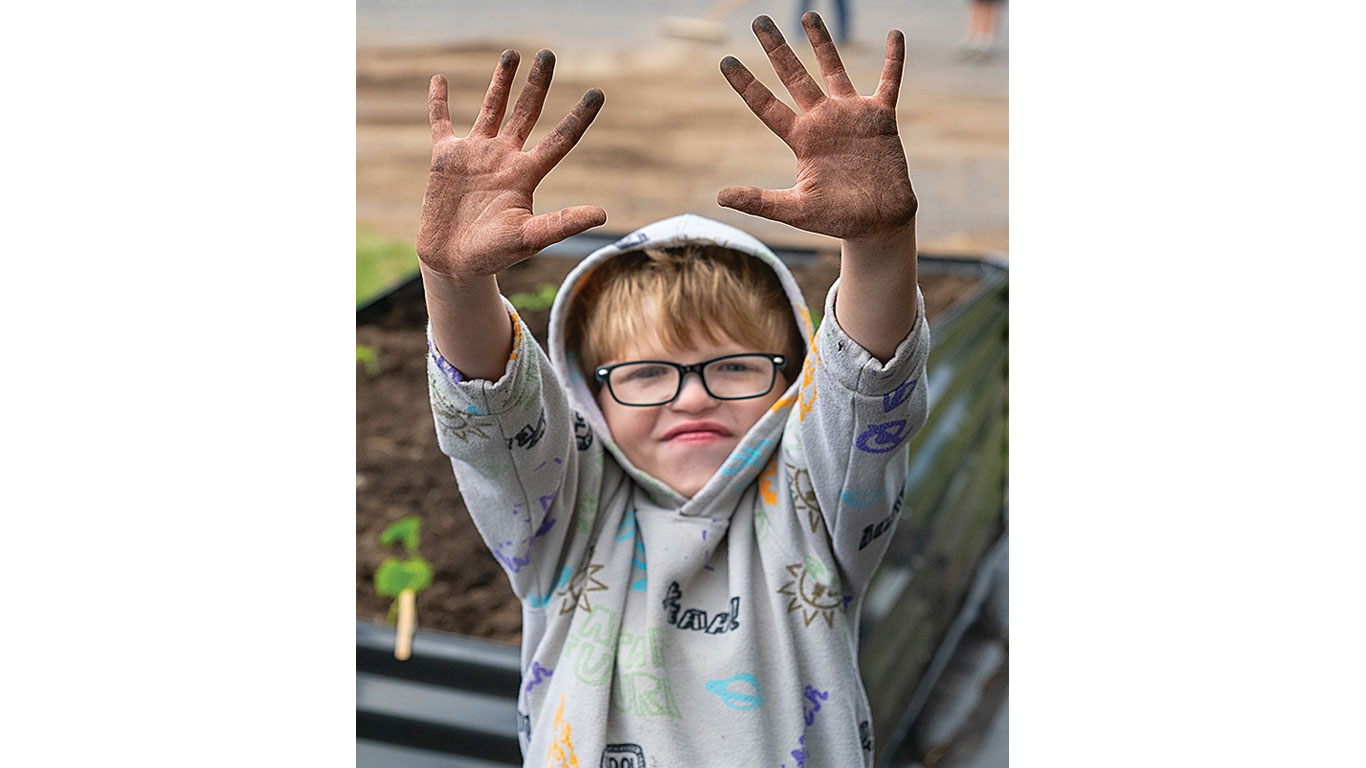 Child in patterned hoodie shows hands covered in dirt near a raised garden bed.