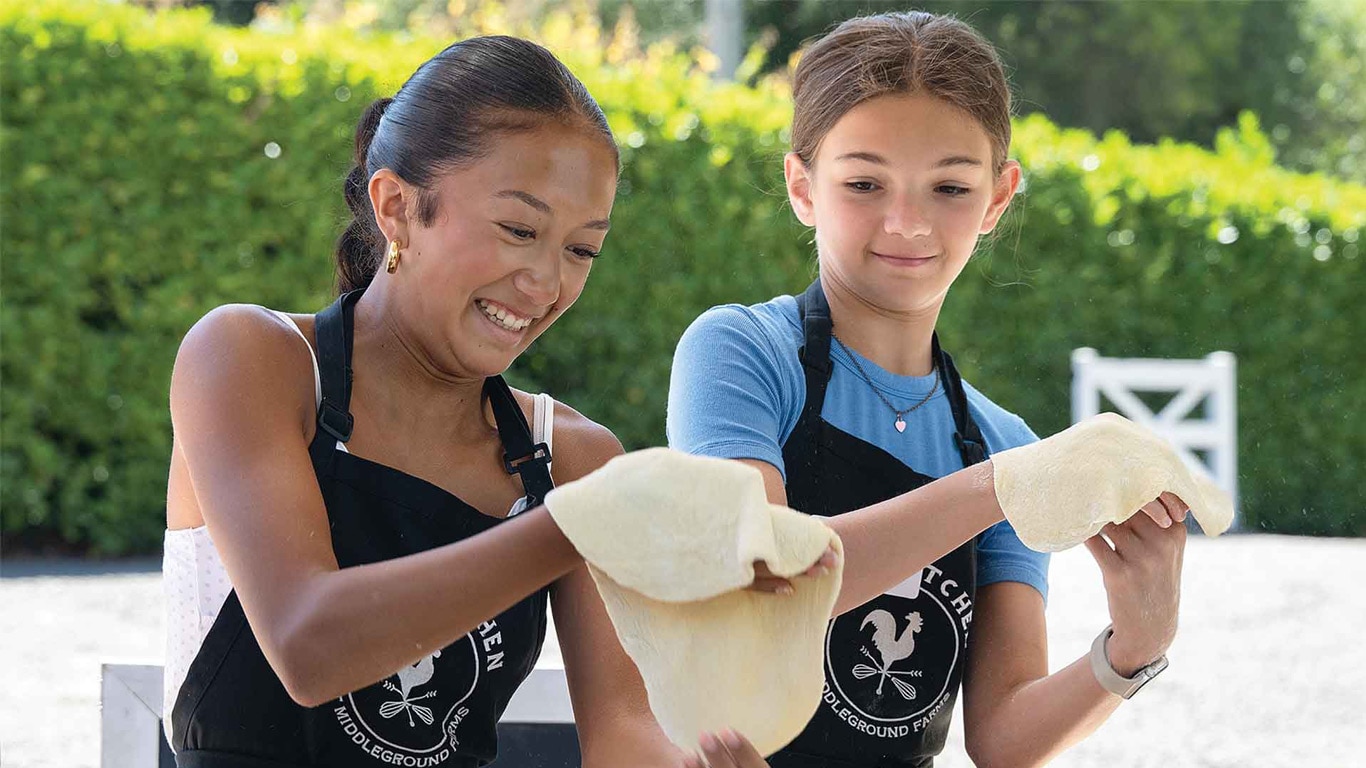 Two children stretching pizza dough outdoors.