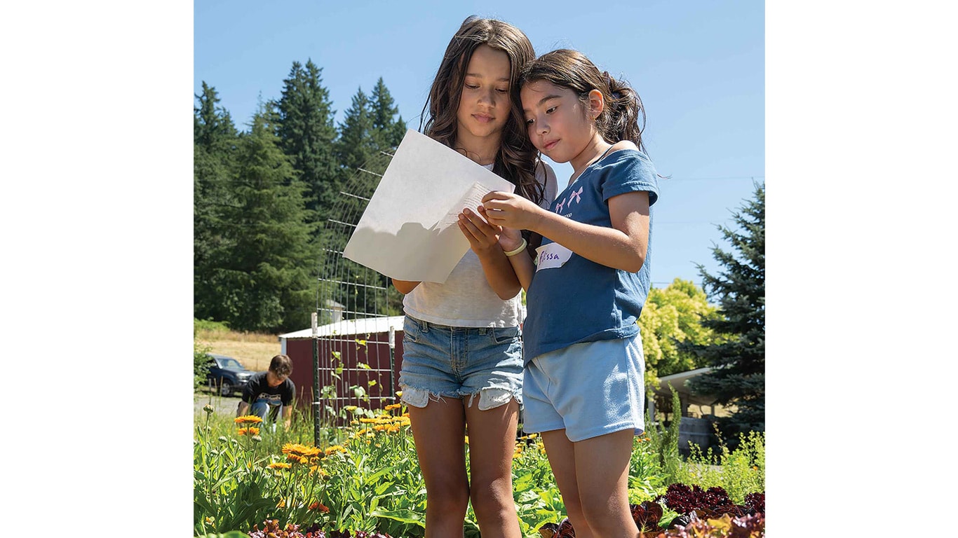 Two children standing outdoors looking at a sheet of paper in a garden.