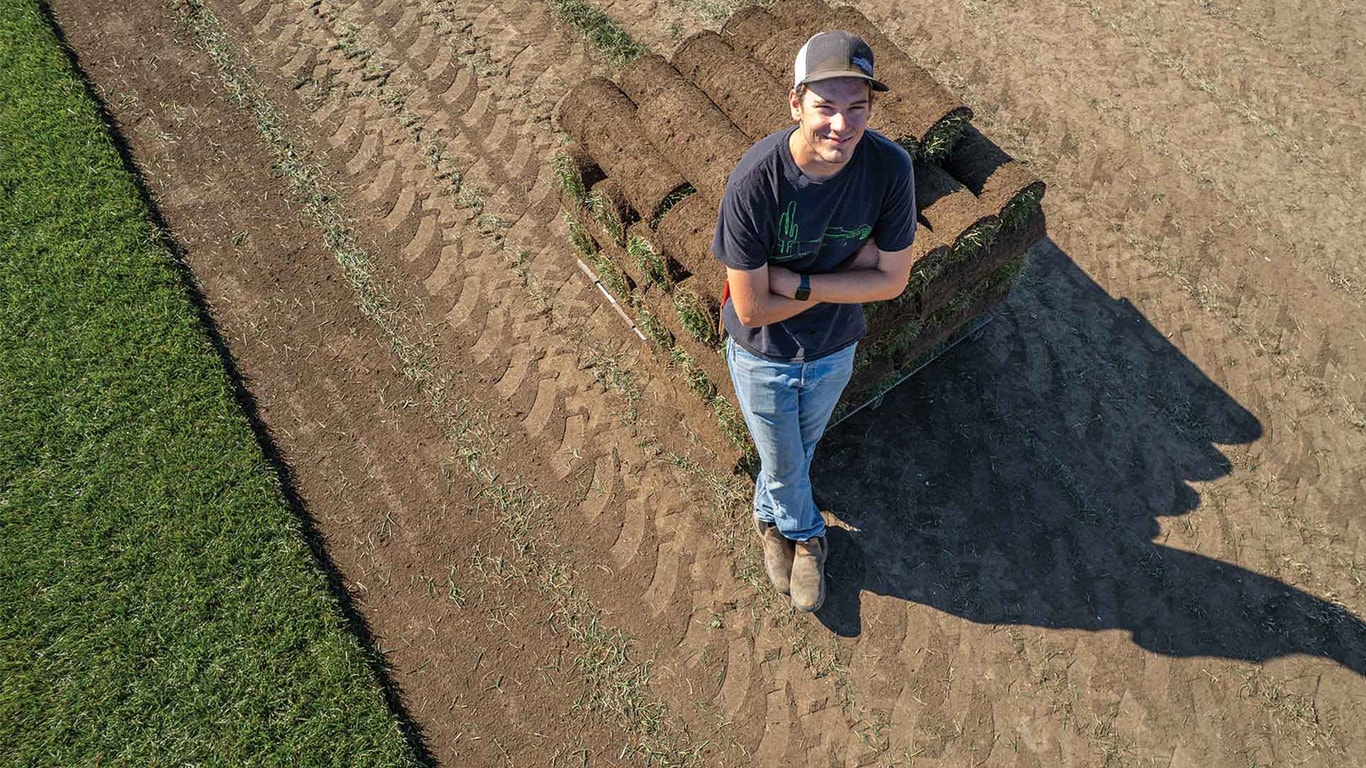Person standing beside a stack of rolled sod on a dirt field.