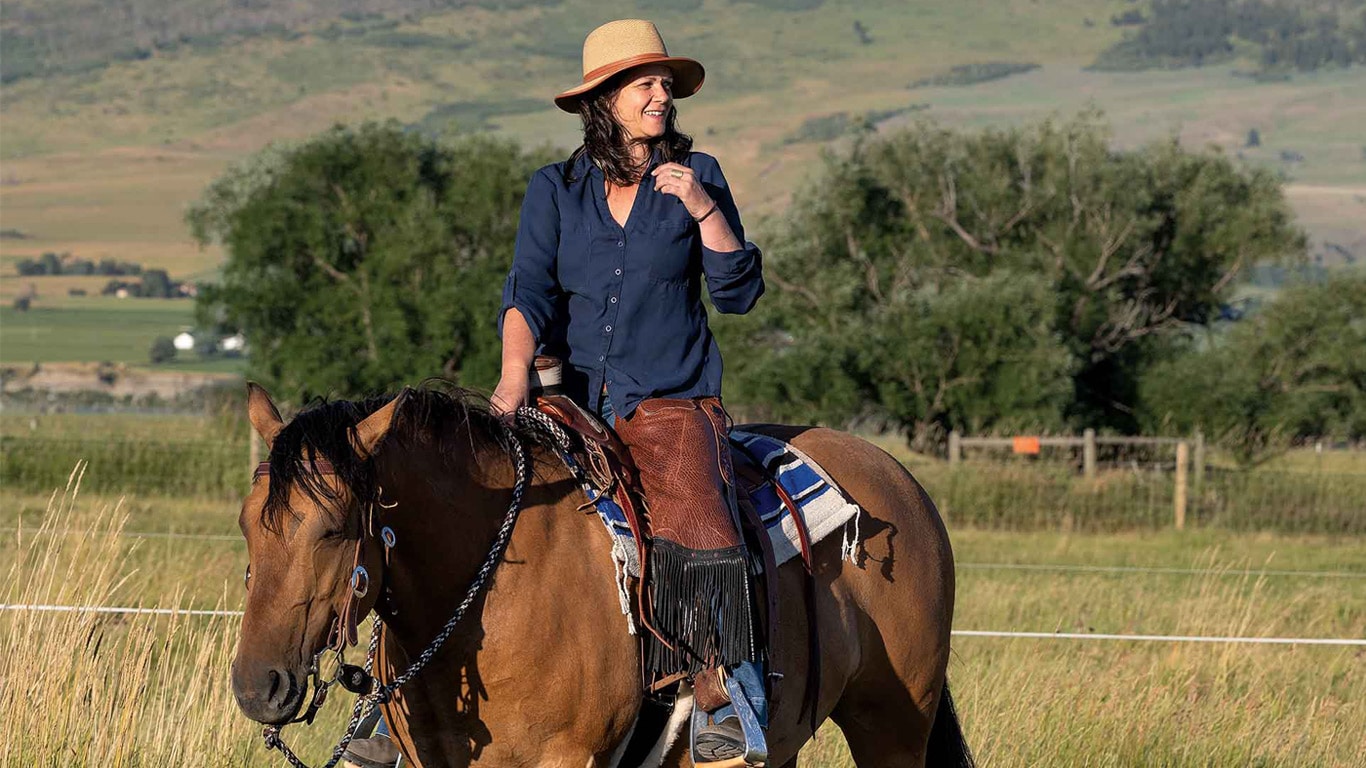 A person riding a horse through tall grass with a mountain range and open fields.