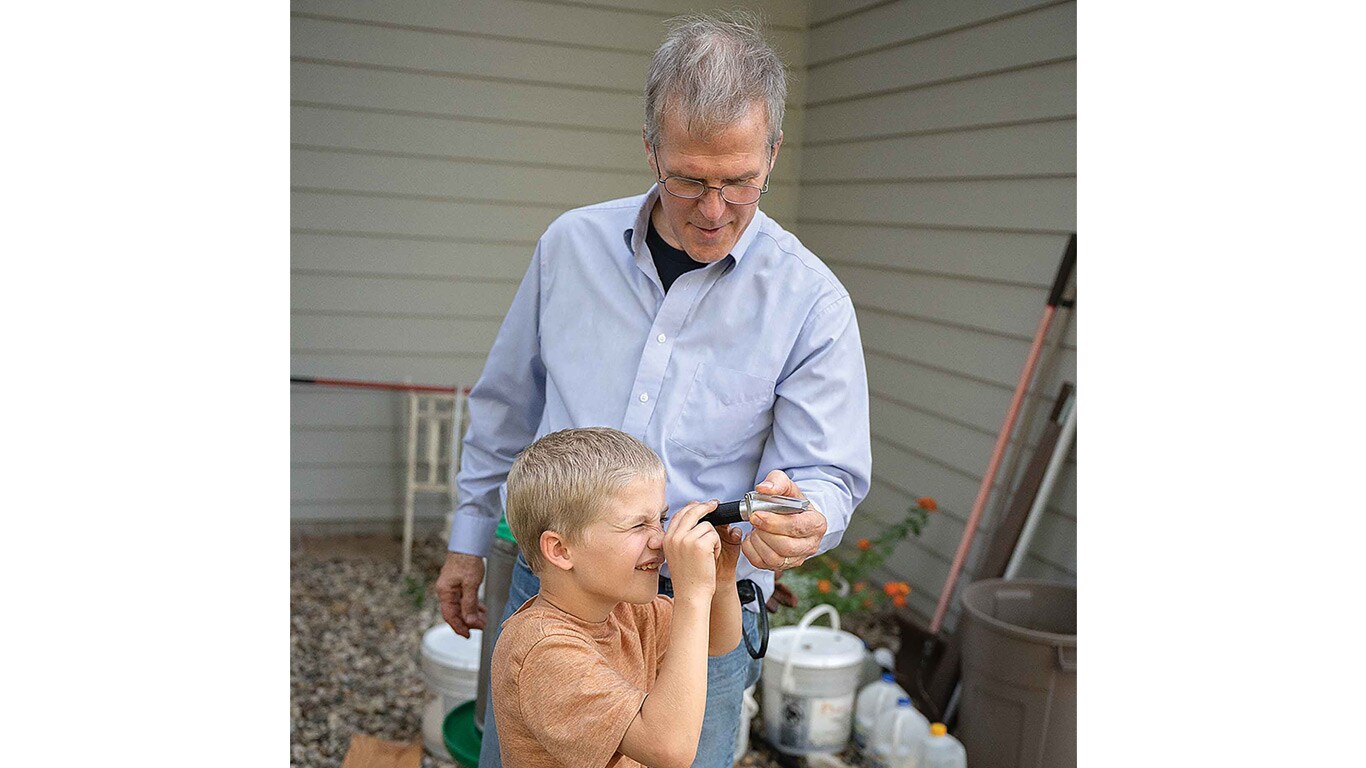 Adult guiding a child using a small handheld tool outdoors beside a house and yard supplies.