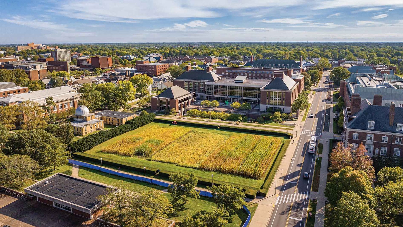 Vue aérienne d'un campus universitaire avec des bâtiments d'enseignement, une rue et un champ de culture entouré d'arbres. 