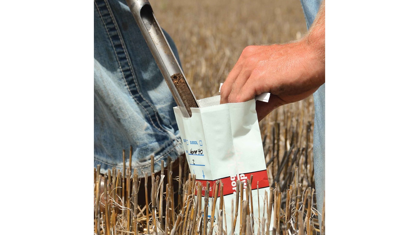 A close-up of a soil sampling tool depositing soil into a paper sample bag.