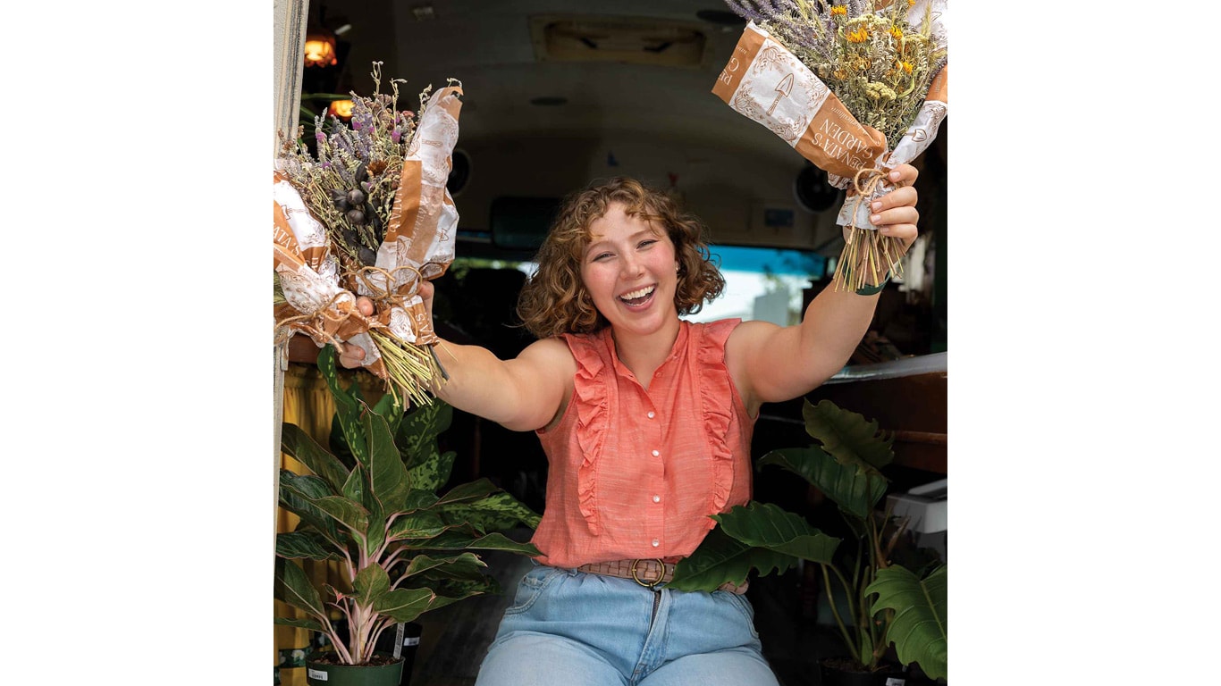 Person sitting in a vehicle doorway holding two wrapped flower bouquets, with potted plants arranged beside them.