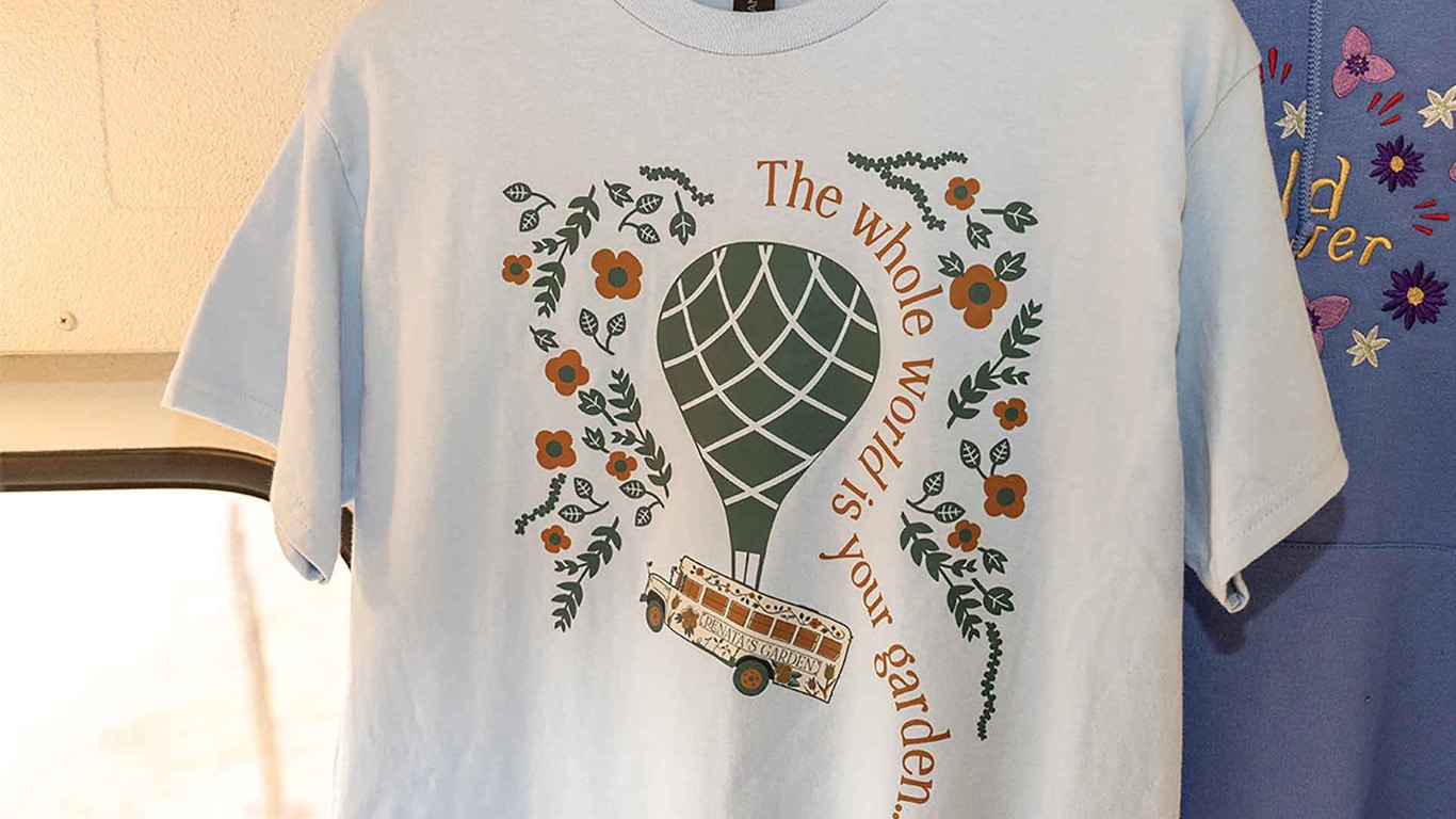 White t-shirt with hot air balloon graphic, flowers, and text reading 'The whole world is your garden.'
