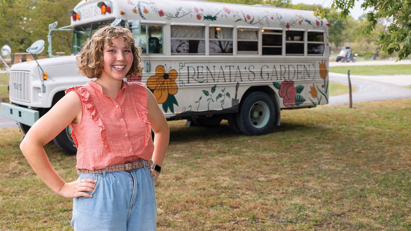 Person standing with hands on hips in front of a floral-painted short bus labeled 'Renata's Garden'.
