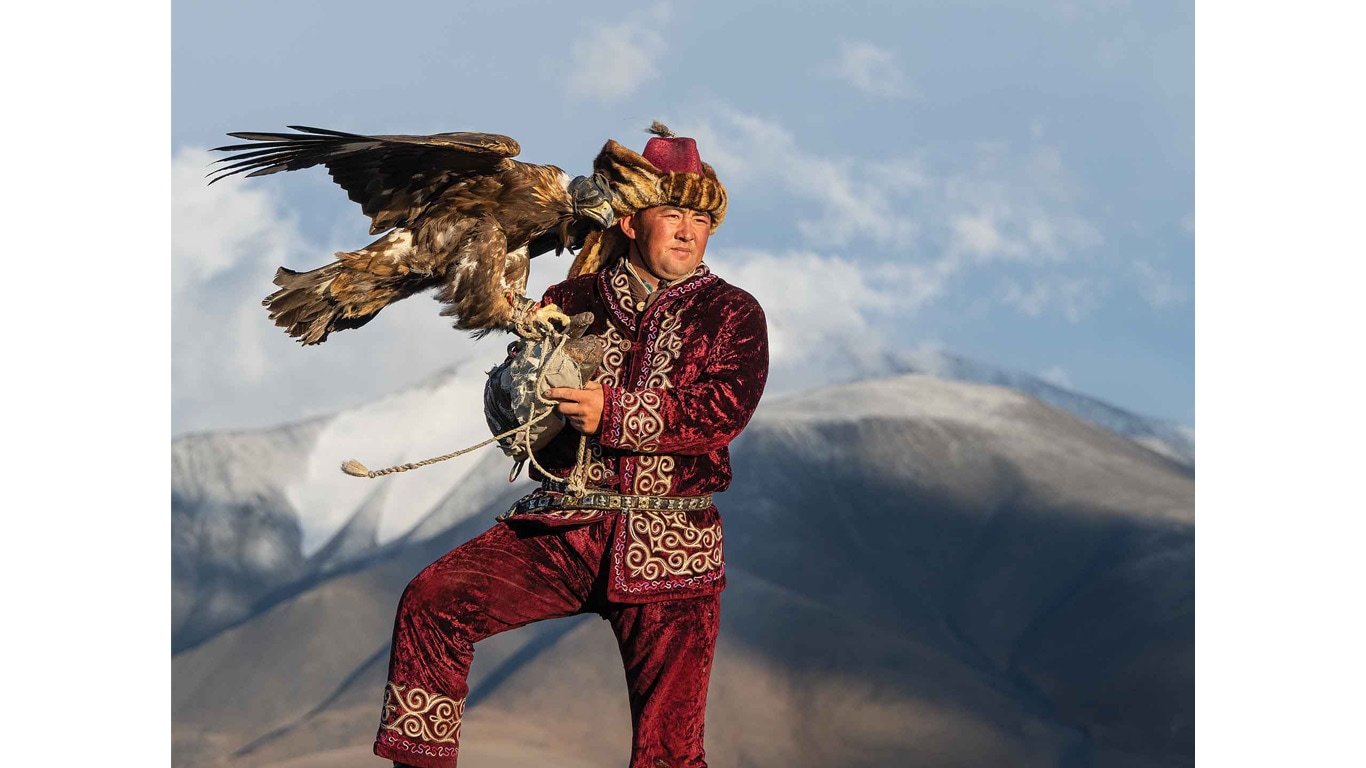 A person in ornate traditional clothing standing on rocky terrain, holding an eagle.