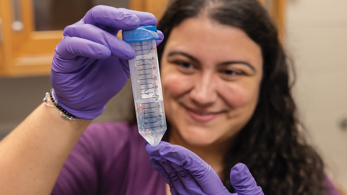 Mains gantées tenant un cylindre gradué contenant des filaments bleus dans un environnement de laboratoire.