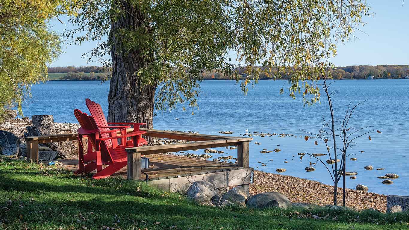 Red Adirondack chairs on a wooden deck beneath a tree overlooking a calm lakeshore.