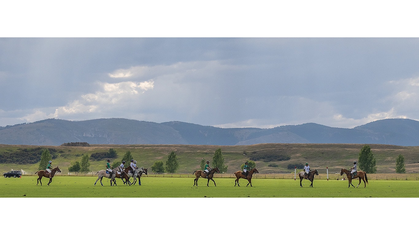 Several riders on horseback spread across a large grassy field with mountains and cloudy sky in the background.