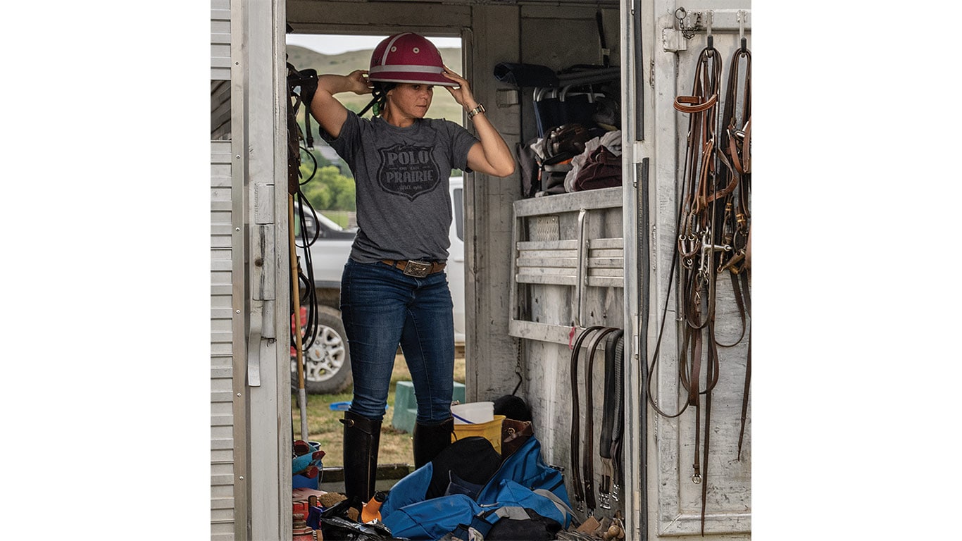 Person inside a tack room adjusting a helmet, with riding gear hanging on the walls.