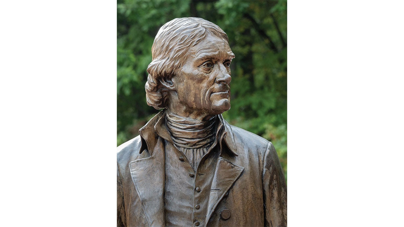 Bronze statue of a historical figure in period clothing, set outdoors with trees in the background.