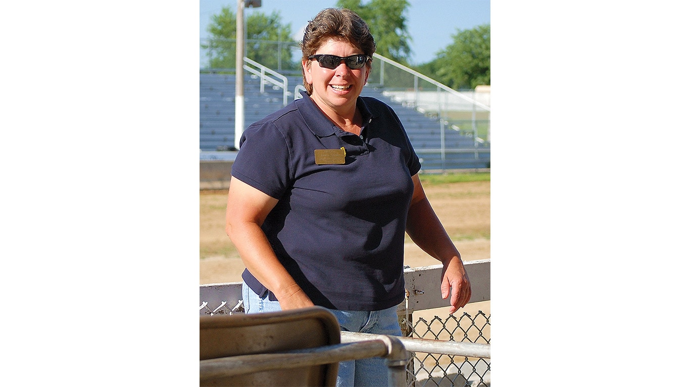 Person wearing sunglasses standing by a fence at an outdoor facility with bleachers in the background.