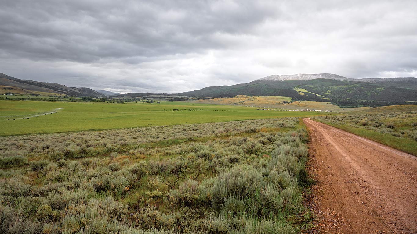 Chemin de terre passant dans une vallée herbeuse en direction de montagnes au loin sous un ciel nuageux.