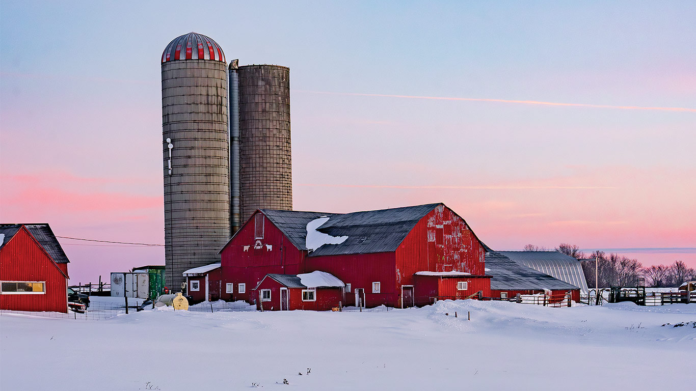 Red barns and tall silos stand in a snowy farmyard at sunset with pink sky in background.
