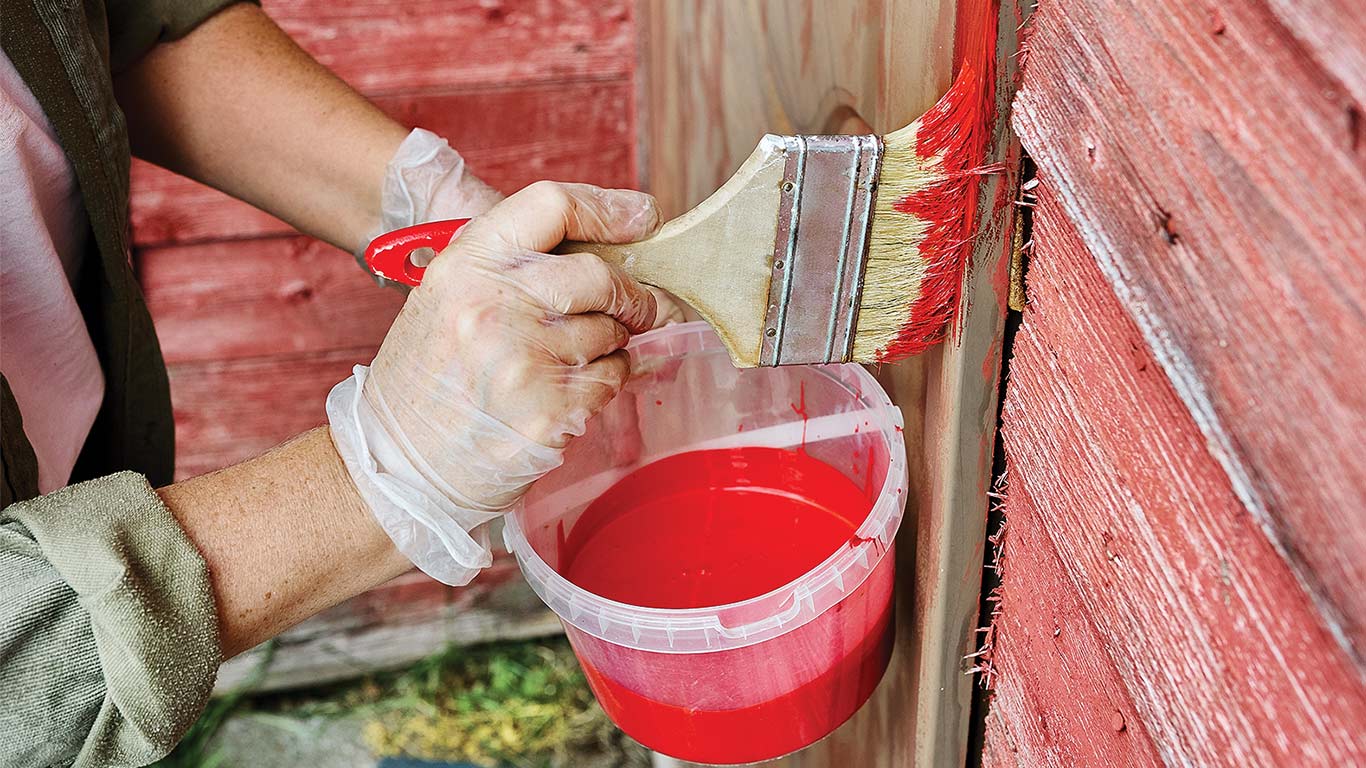 Hands using a paintbrush to apply red paint to a wooden surface.