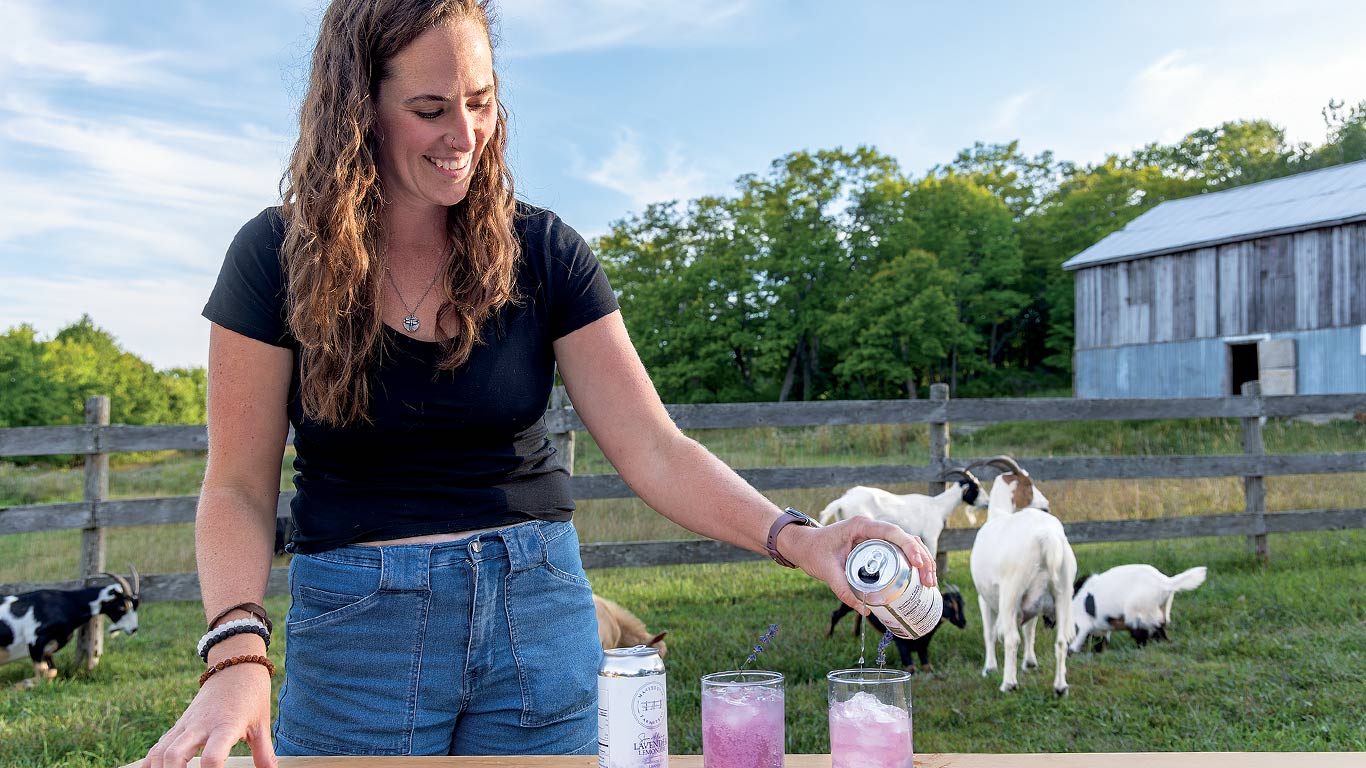 Personne qui verse une boisson à la lavande à l'extérieur avec des chèvres et une grange derrière.