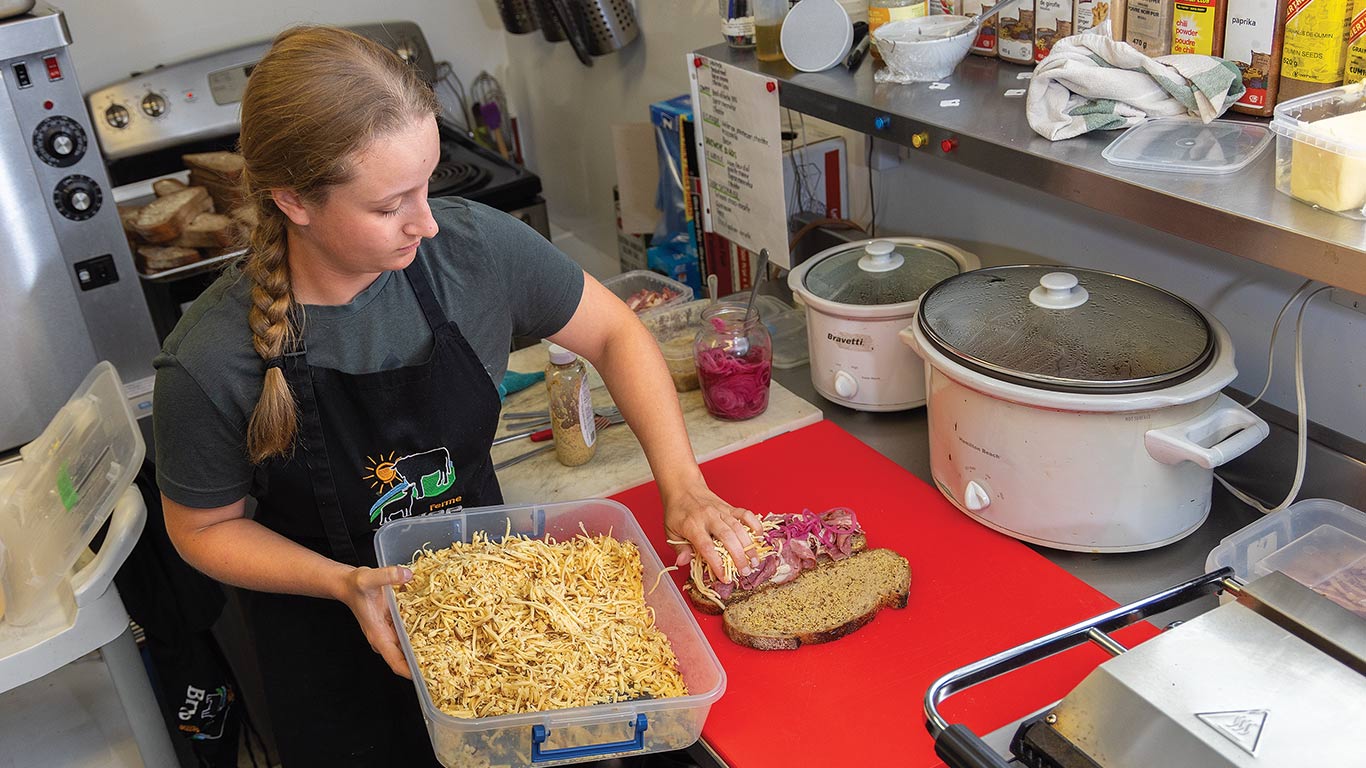 Personne qui prépare un sandwich dans une cuisine, en ajoutant une garniture râpée sur le pain, à côté de mijoteuses.